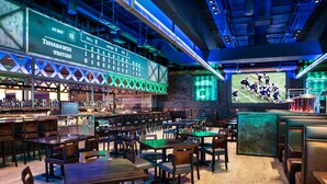 Eine Sportsbar mit mehreren Tischen und Stühlen, einem großen Bildschirm, der ein Fußballspiel zeigt, und einer Anzeigetafel.