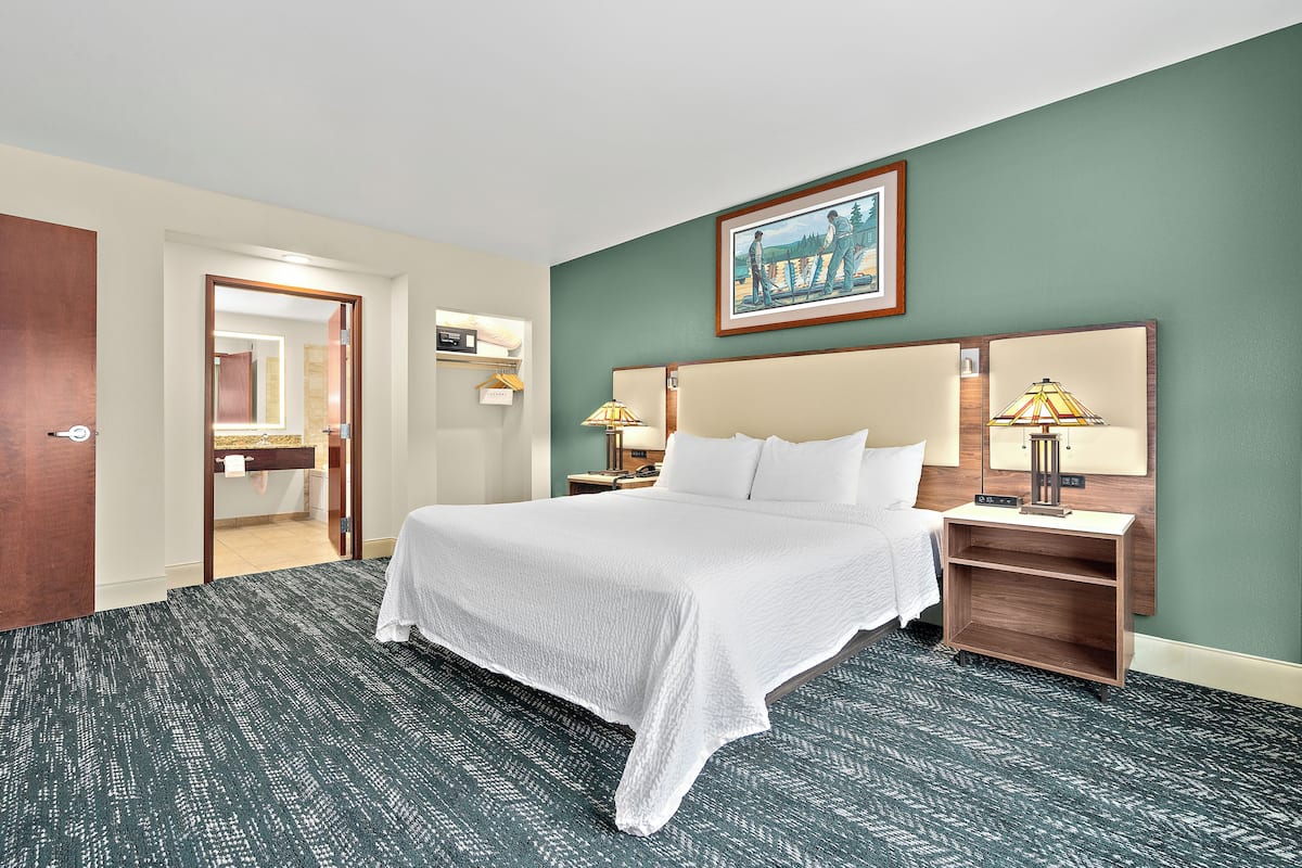 Habitación de hotel con una cama grande, una mesita de noche con lámpara, cabecero de madera, un cuadro en la pared y un baño visible a través de una puerta abierta.
