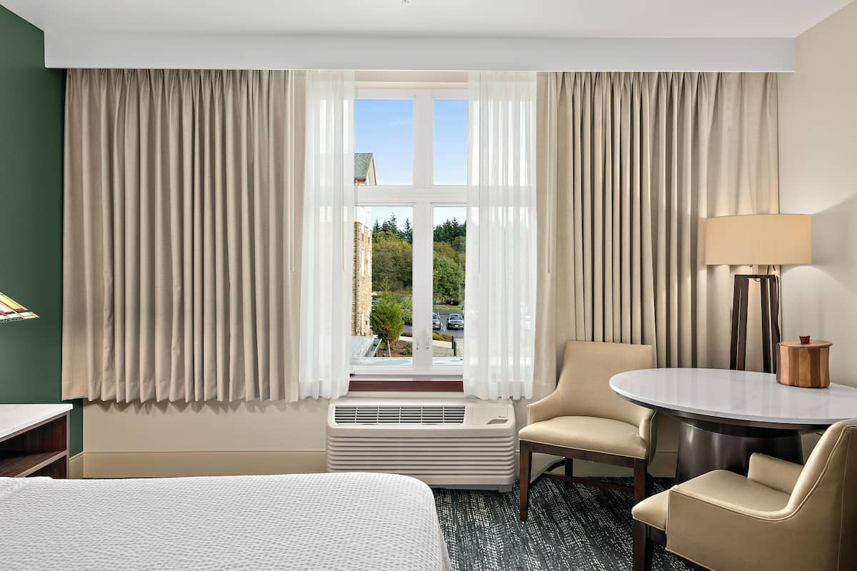 Habitación de hotel con cama, una mesa redonda, dos sillas, un escritorio, una lámpara y un ventanal con cortinas.