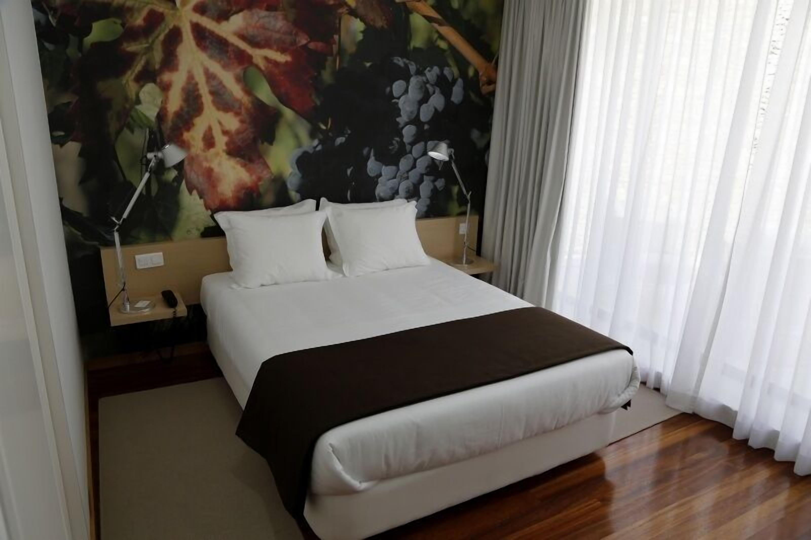Habitación de hotel con una cama grande, ropa de cama blanca, una manta oscura, una mesita de noche con una lámpara y un mural en la pared con uvas y hojas.