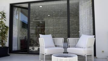 A modern outdoor seating area with two white wicker chairs and a round table, adjacent to a large glass sliding door.