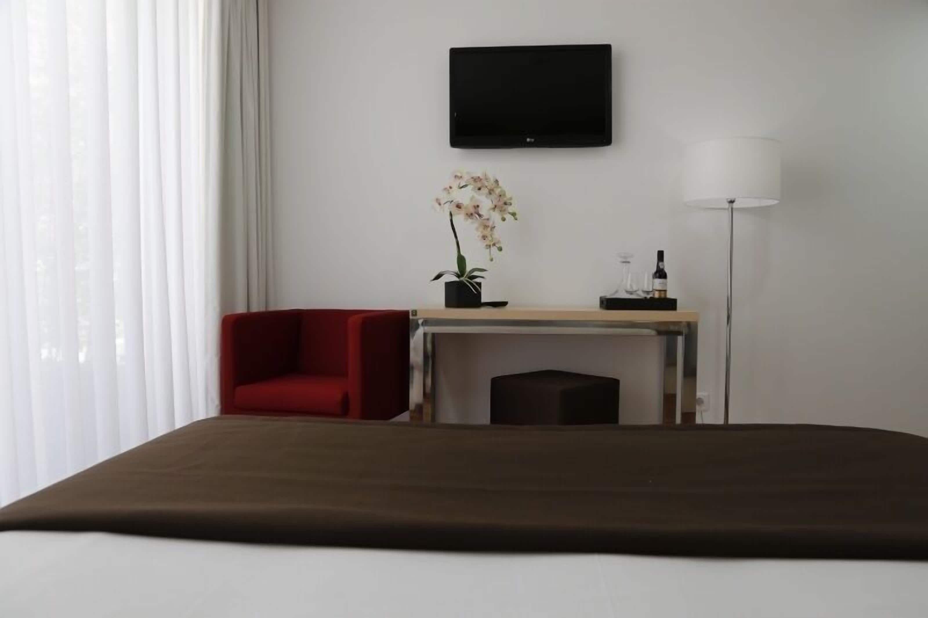A hotel room with a bed, a red chair, a desk with a plant, a TV, and a lamp.