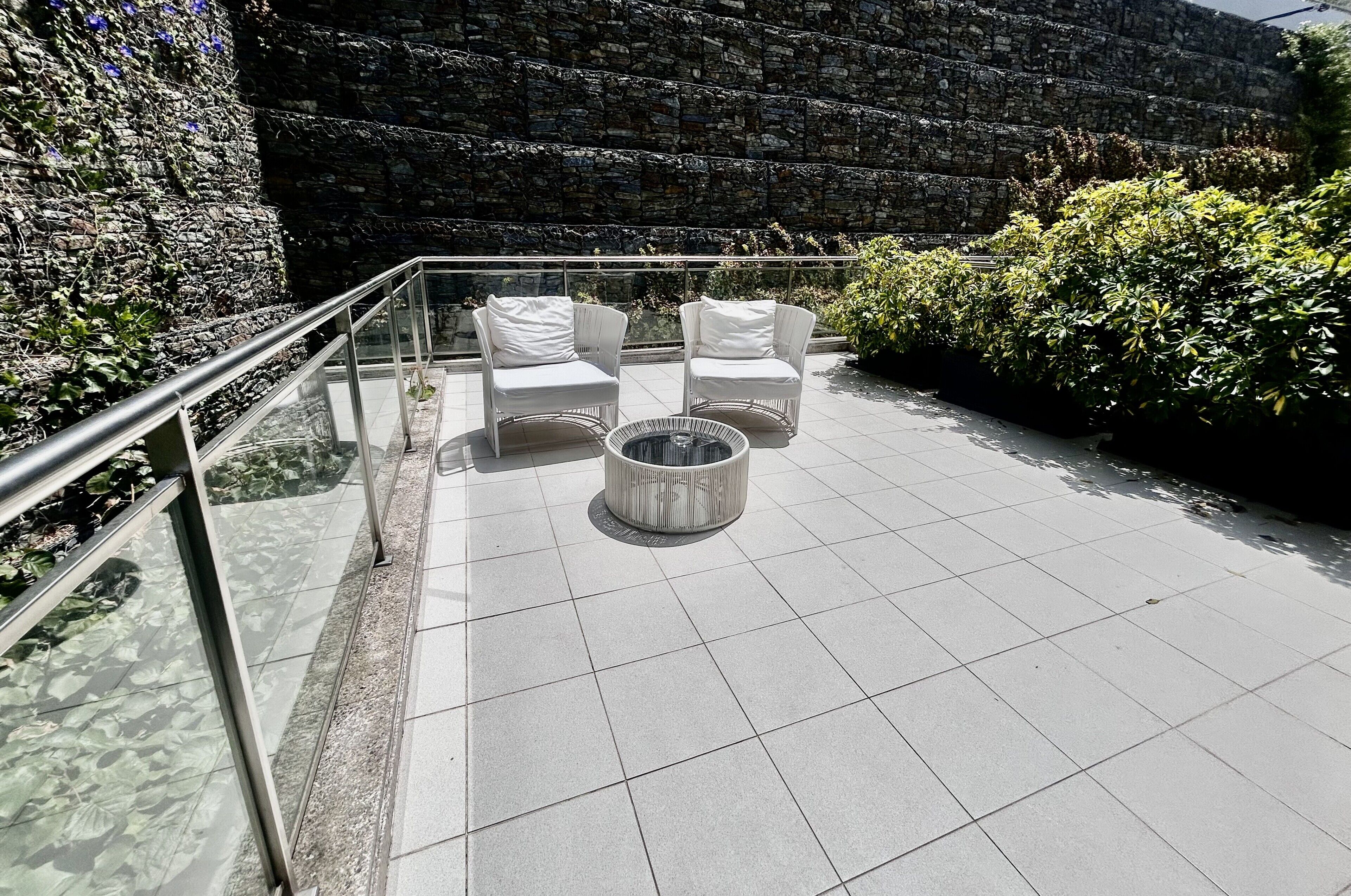 Una terrazza sul tetto con due poltrone bianche imbottite e un tavolino rotondo, circondata da un muro in pietra e vegetazione.