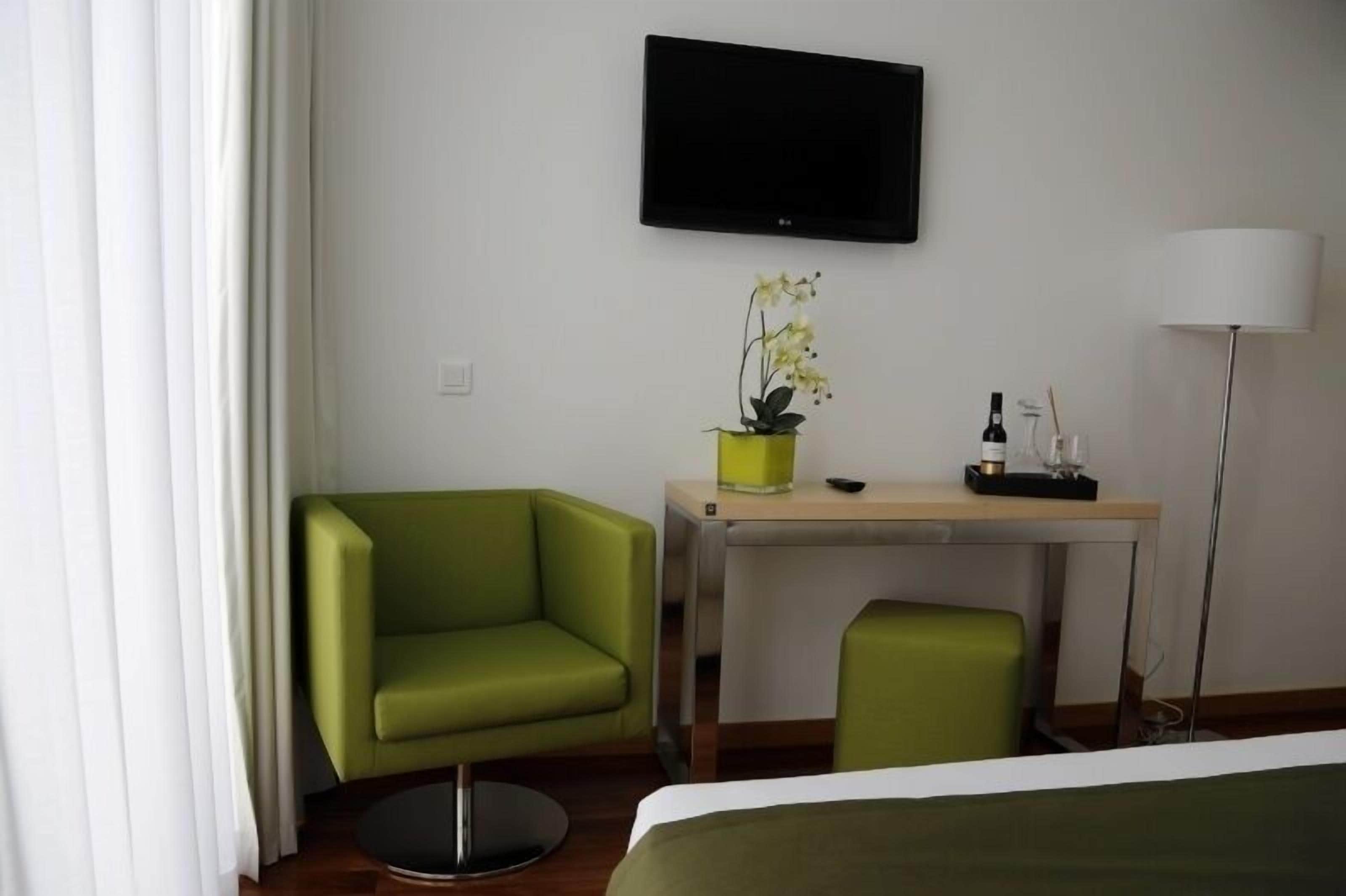Et hotelværelse med en grøn lænestol, et lille skrivebord med en orkidé i potte, et fladskærms-tv og en sengebordlampe.
