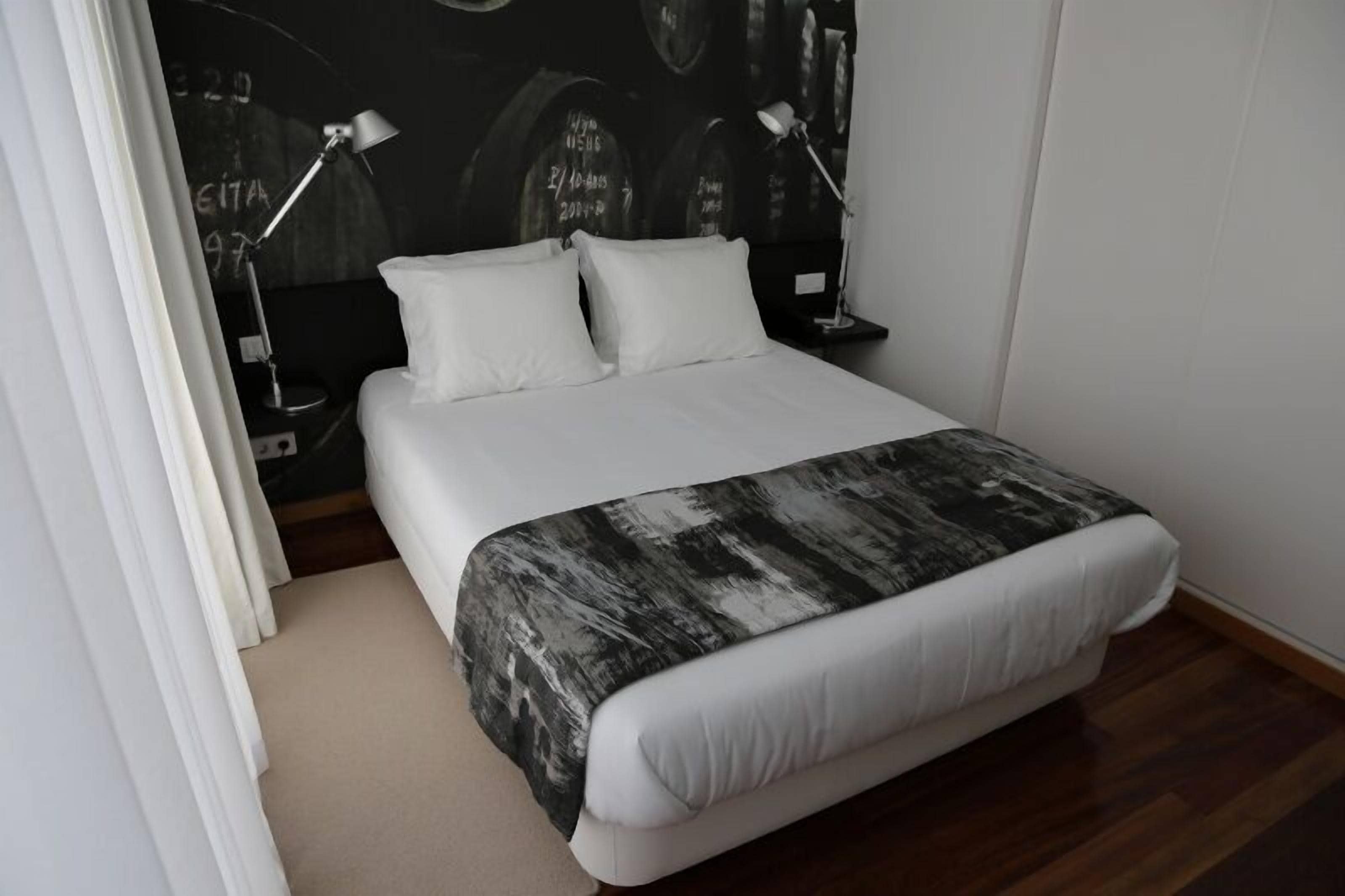 A neatly made bed with white linens and a black and white patterned throw, two bedside lamps, and a wall with abstract designs.