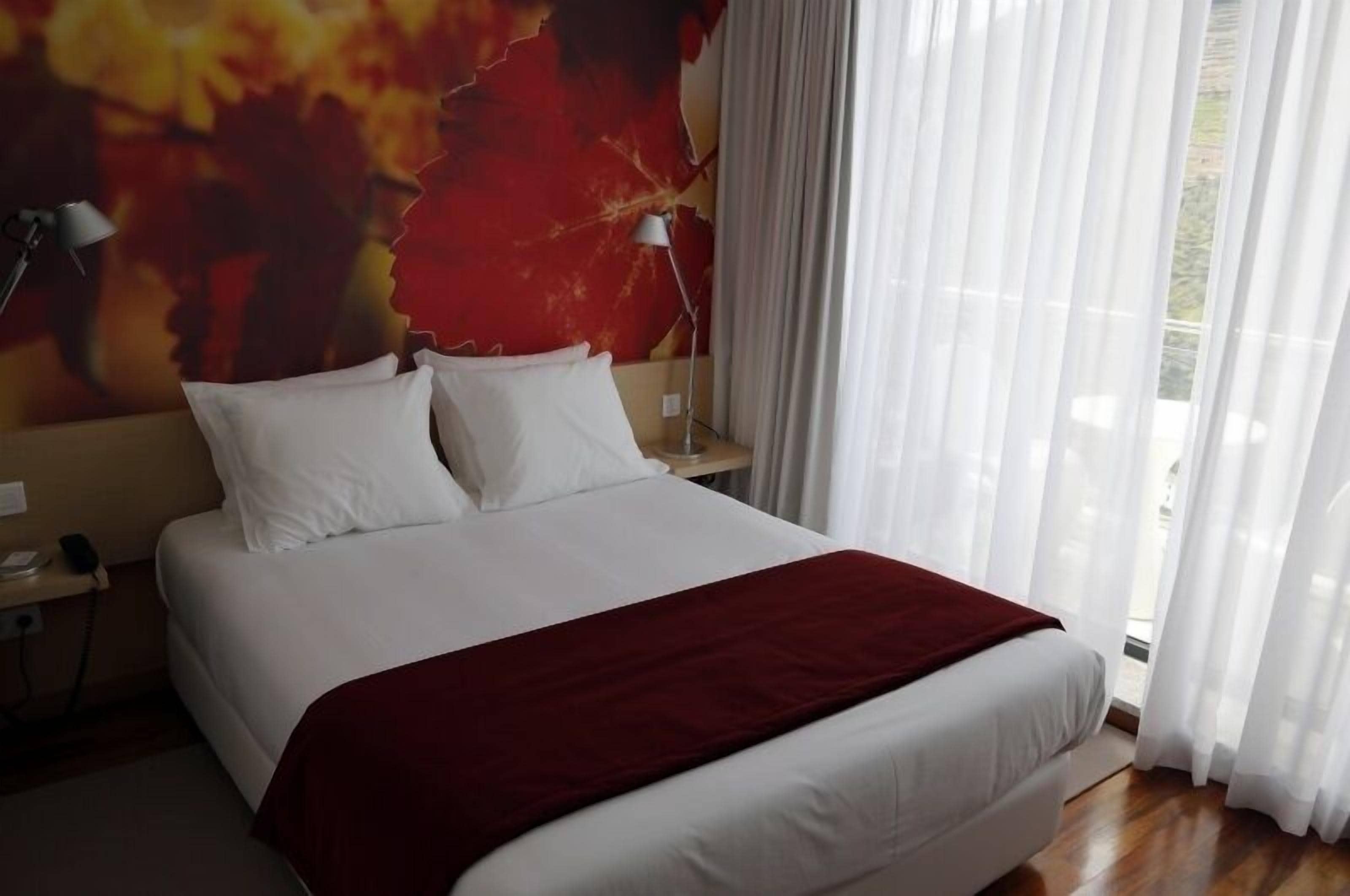 A neatly made bed with white linens and a red throw blanket, a bedside lamp, and a wall with a large abstract painting.