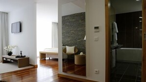Una sala de estar moderna con un espejo grande, piso de madera, un sofá, una silla y una pequeña mesa con un jarrón de flores.