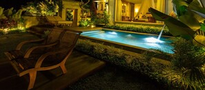 A nighttime pool area with two wooden lounge chairs, a small waterfall feature, and lush tropical plants. - Jiwa Klusa Luxury Villa (Payangan)