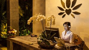 Een receptiebalie met een laptop, een telefoon en een decoratief wandpaneel. Voor op de foto staan potplanten en een houten tafel.