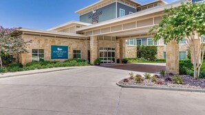 L’entrée d’un hôtel, avec une enseigne indiquant « Homewood Suites Hilton », se caractérise par une façade en pierre, une entrée abritée et des espaces paysagers agrémentés de plantes.