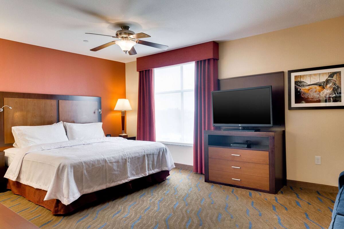 Une chambre d’hôtel équipée d’un lit, d’une télévision, d’un bureau, d’une lampe, d’un ventilateur de plafond et d’une fenêtre avec des rideaux.