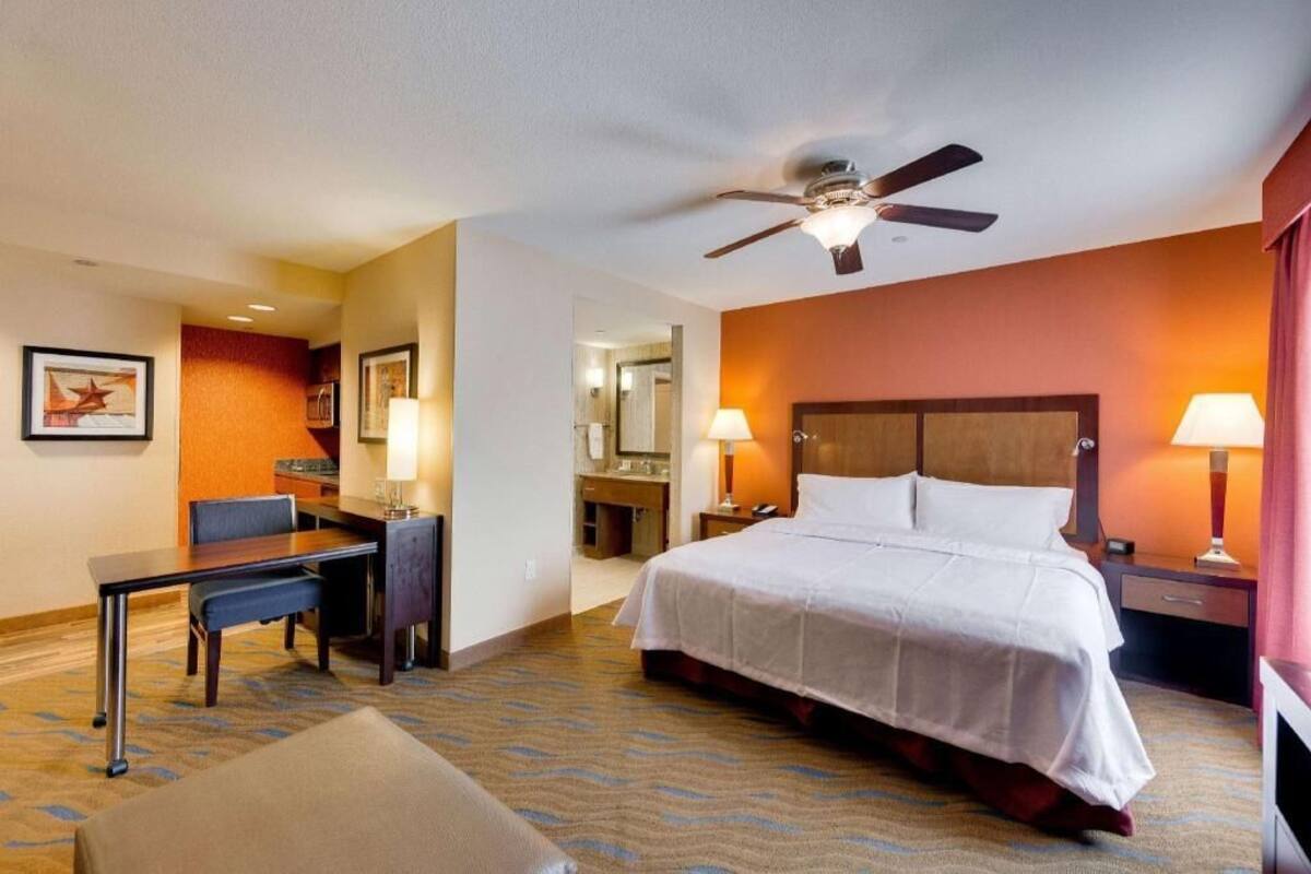 Une chambre d’hôtel équipée d’un lit, d’un bureau, d’une chaise, d’un ventilateur de plafond et de deux lampes de chevet.