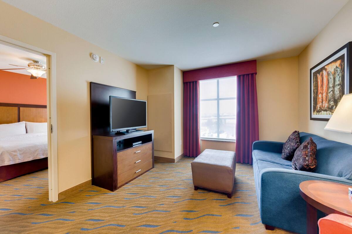 Une chambre d’hôtel avec un lit, un téléviseur posé sur un meuble en bois, un canapé bleu, une petite table et une fenêtre avec des rideaux.