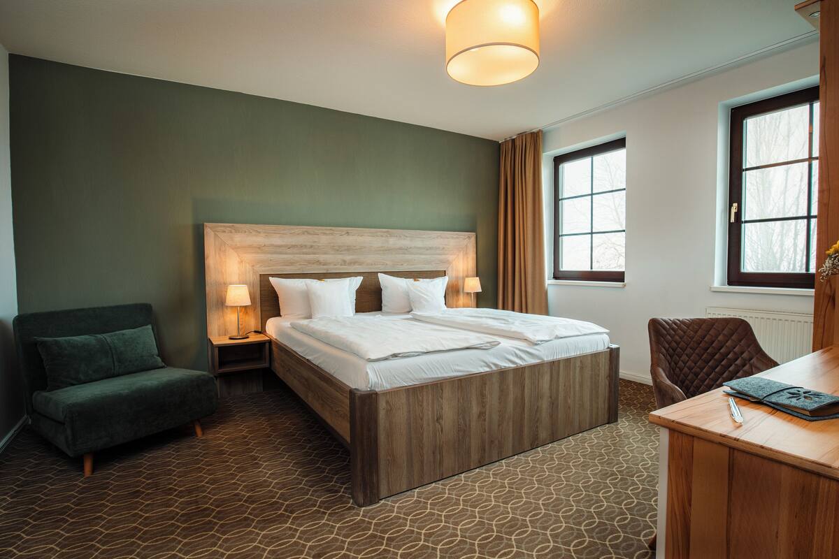 A hotel room with a large bed, a desk with a chair, a green armchair, and a window with curtains.