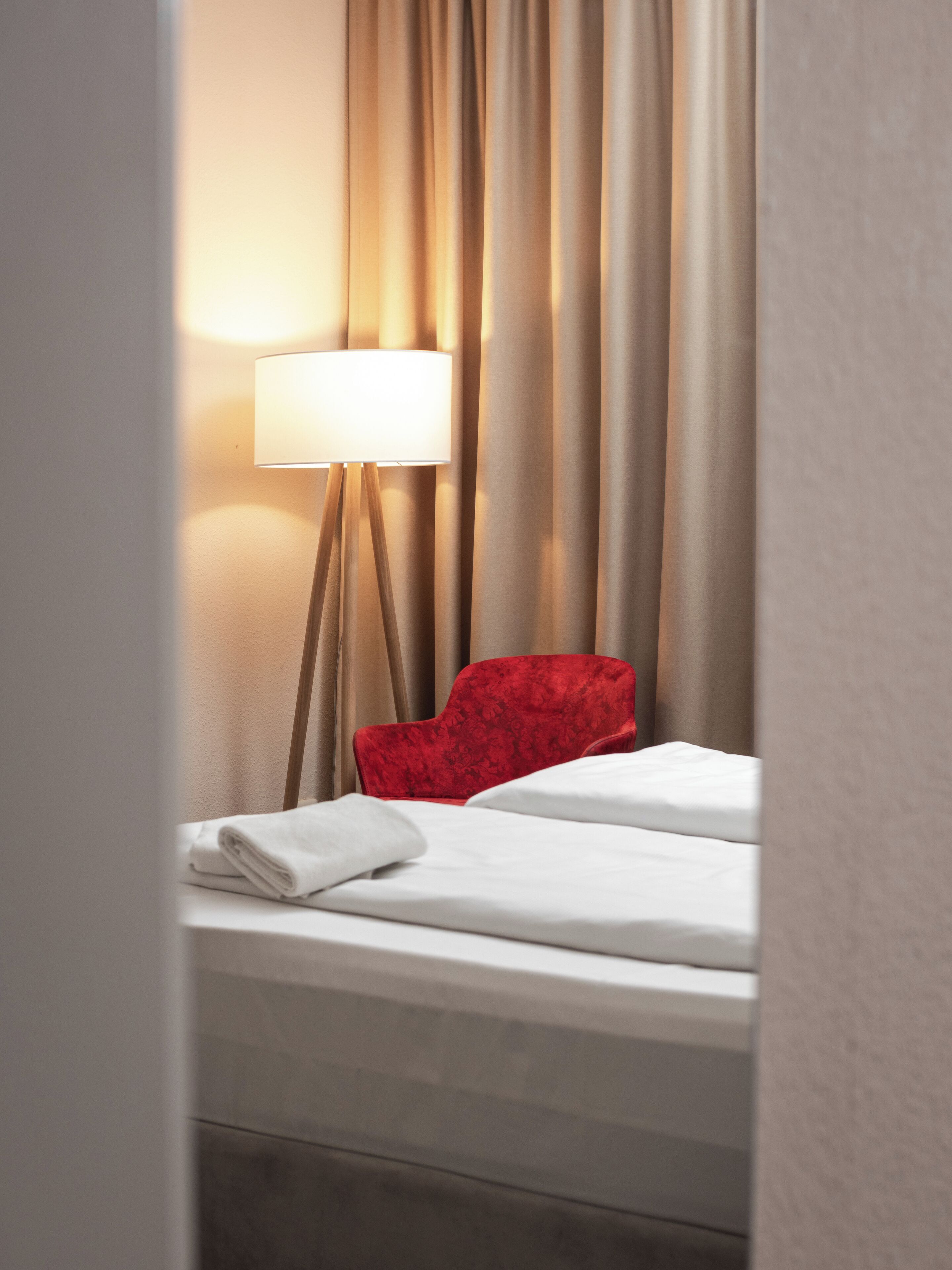 Una habitación de hotel con cama, una silla roja, una lámpara de pie y cortinas.