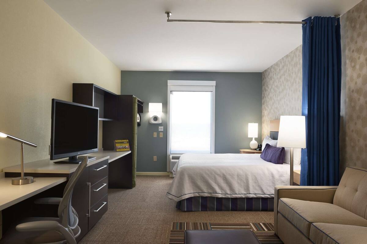 Une chambre d’hôtel comprenant un lit, un bureau avec un ordinateur, une chaise, une télévision, une lampe et une fenêtre avec des rideaux.