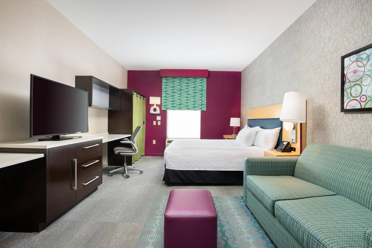 Une chambre d’hôtel avec un lit, un bureau doté d’un téléviseur à écran plat, un canapé vert et une petite table avec une lampe.