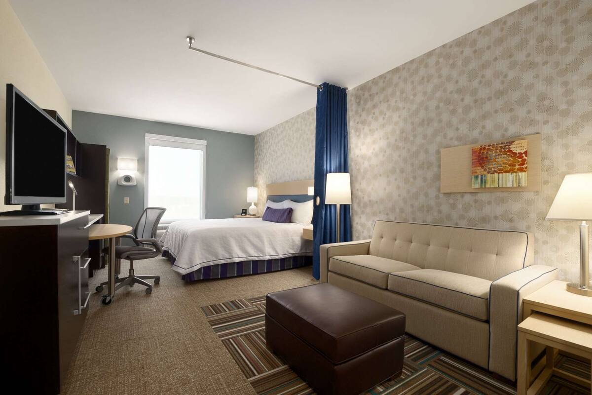 Une chambre d’hôtel avec un lit, un canapé, un bureau avec un ordinateur, une télévision et une fenêtre avec des rideaux.