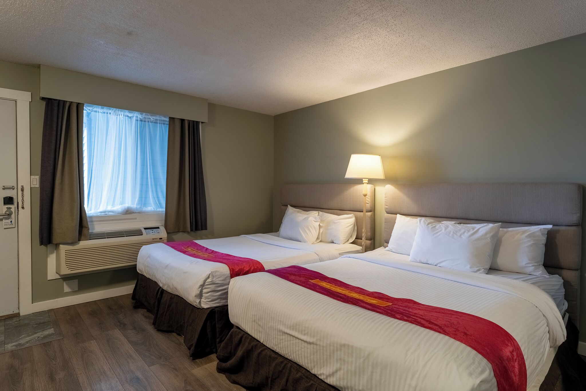 A hotel room with two beds, a window with curtains, an air conditioning unit, and a bedside lamp.