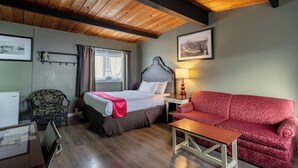 Une chambre d’hôtel avec un plafond en bois, un lit doté d’une tête de lit grise, un canapé rouge à motifs, une petite table, une chaise et une lampe.
