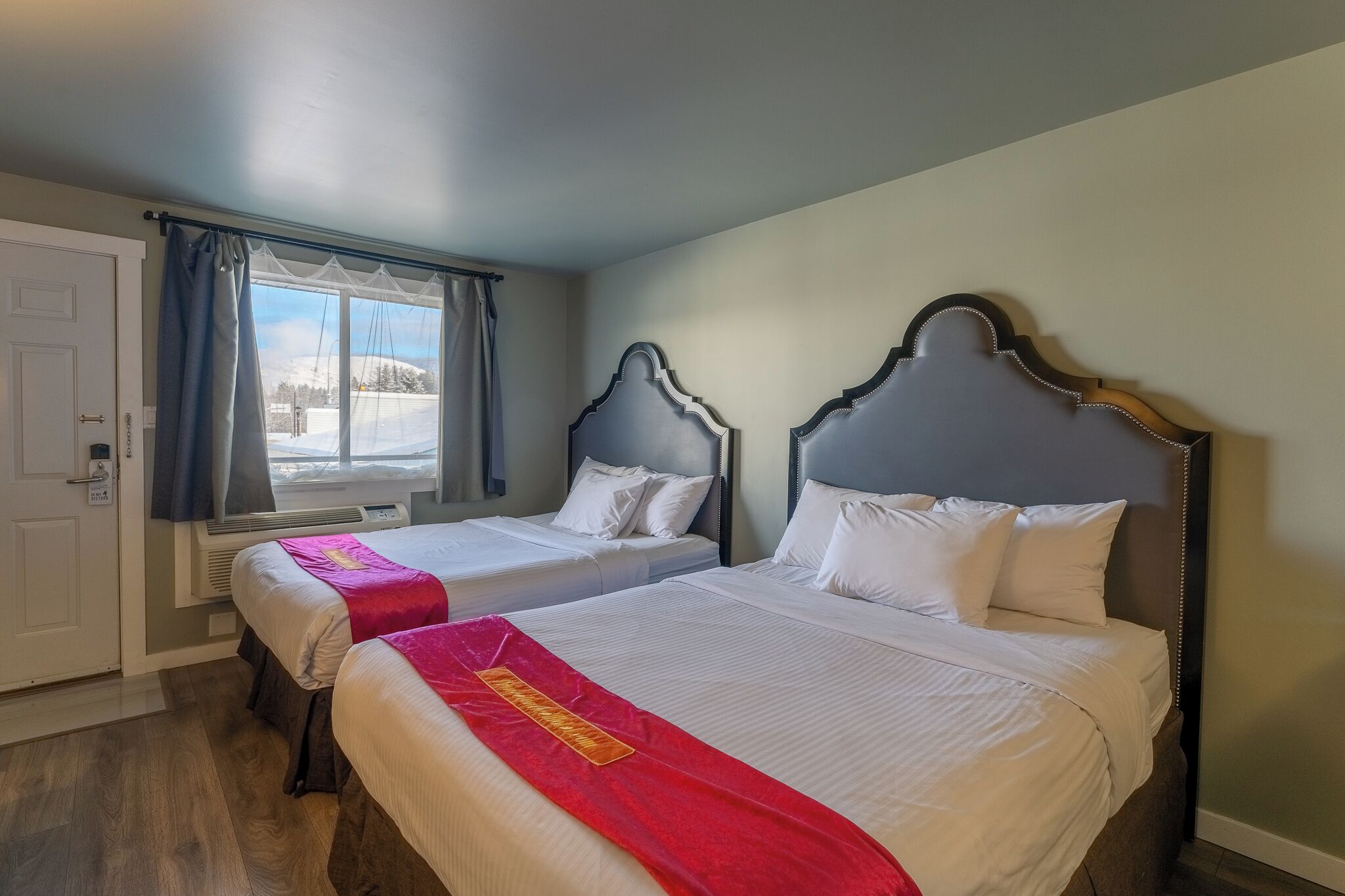 A hotel room with two beds, a window with curtains, a door, and a view of a snowy landscape.