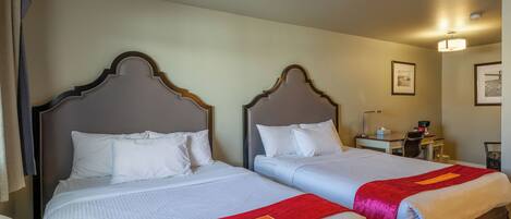 Two beds with wooden headboards, white bedding, and red throw blankets. There is a desk with a chair and a lamp in the room.