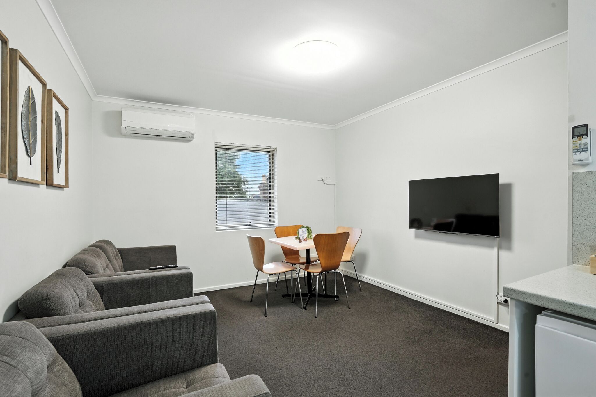 Un salon moderne avec un canapé d’angle gris, une table à manger avec quatre chaises, un téléviseur à écran plat fixé au mur et un appareil de climatisation également fixé au mur.