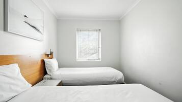 A small, clean room with two beds, a wooden headboard, a window with blinds, and a wall-mounted light fixture.