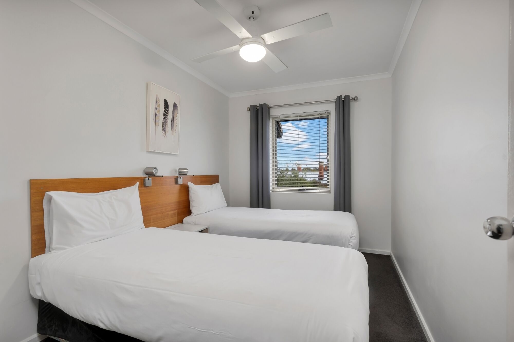 Une chambre d’hôtel avec deux lits simples, un ventilateur de plafond, une fenêtre avec des stores et une œuvre d’art encadrée au mur.