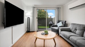 A modern living room with a flat-screen TV, a grey sofa, a round wooden coffee table, and a view of a backyard with a barbecue grill. - Elphin Motel & Serviced Apartments (Launceston)