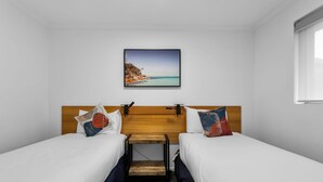 A twin room with two beds, a wooden headboard, a small wooden table, and a painting of a coastal landscape. - Elphin Motel & Serviced Apartments (Launceston)