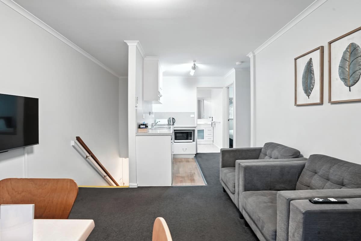 A modern living room with a grey sofa, a wooden chair, and a flat-screen TV. A kitchen is visible in the background with white cabinets and a microwave.