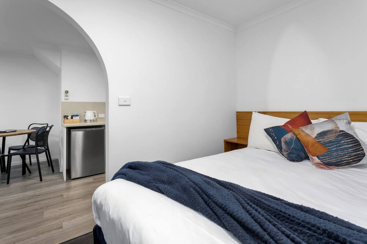 A hotel room with a bed, a desk with a chair, a kitchenette, and a wooden headboard with pillows.