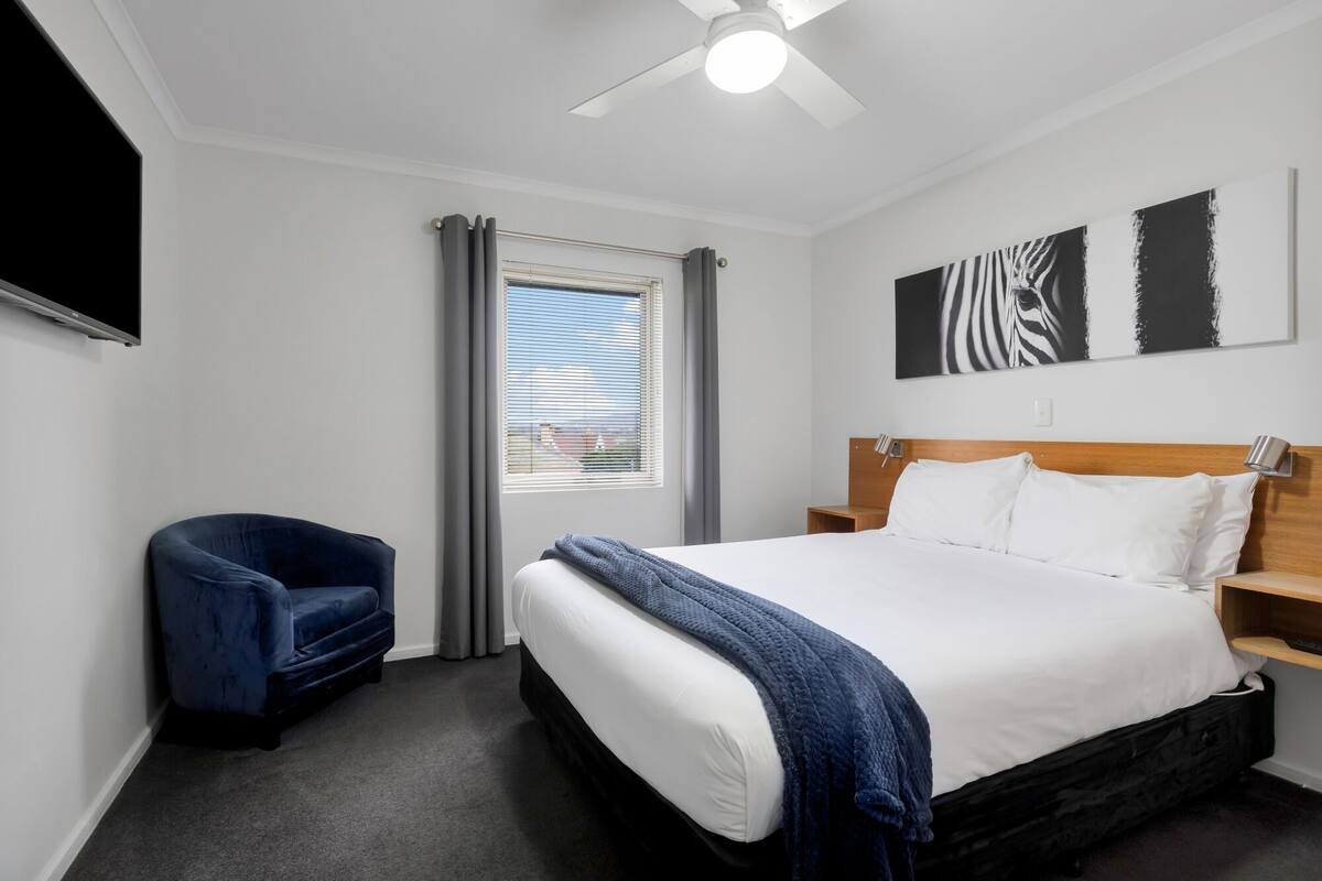 A hotel room with a bed, a chair, a ceiling fan, a window with blinds, and artwork on the wall.