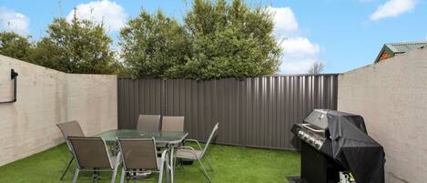 A backyard with a glass table and chairs, a barbecue grill, and a propane tank.