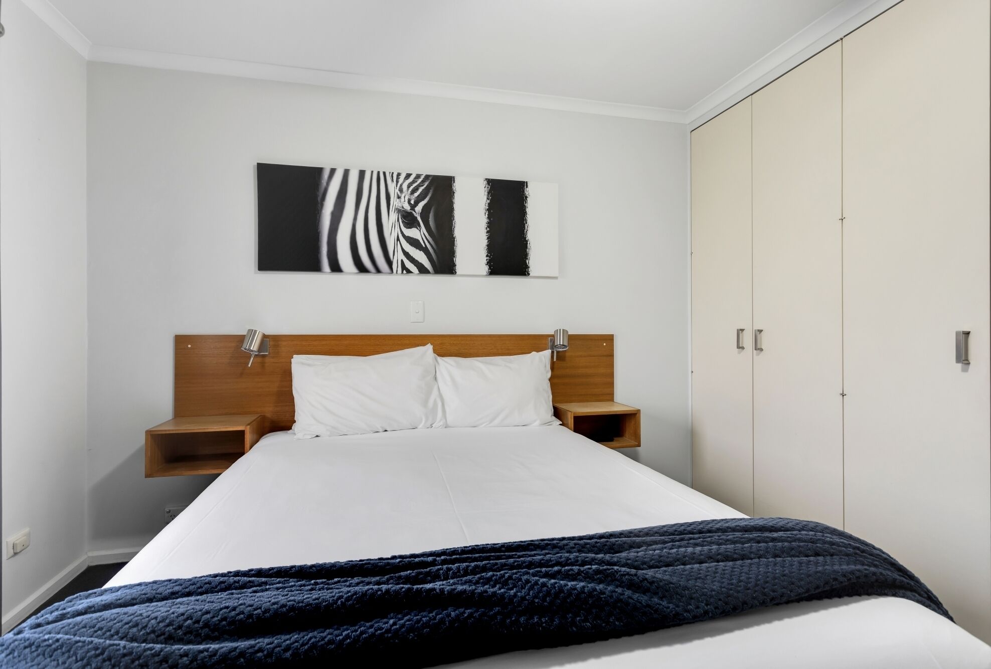 Una camera da letto con un letto, cuscini bianchi, una testiera in legno, un comodino, un'opera d'arte raffigurante una zebra appesa al muro e un armadio.