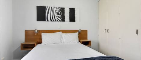 Una camera da letto con un letto, cuscini bianchi, una testiera in legno, un comodino, un'opera d'arte raffigurante una zebra appesa al muro e un armadio.
