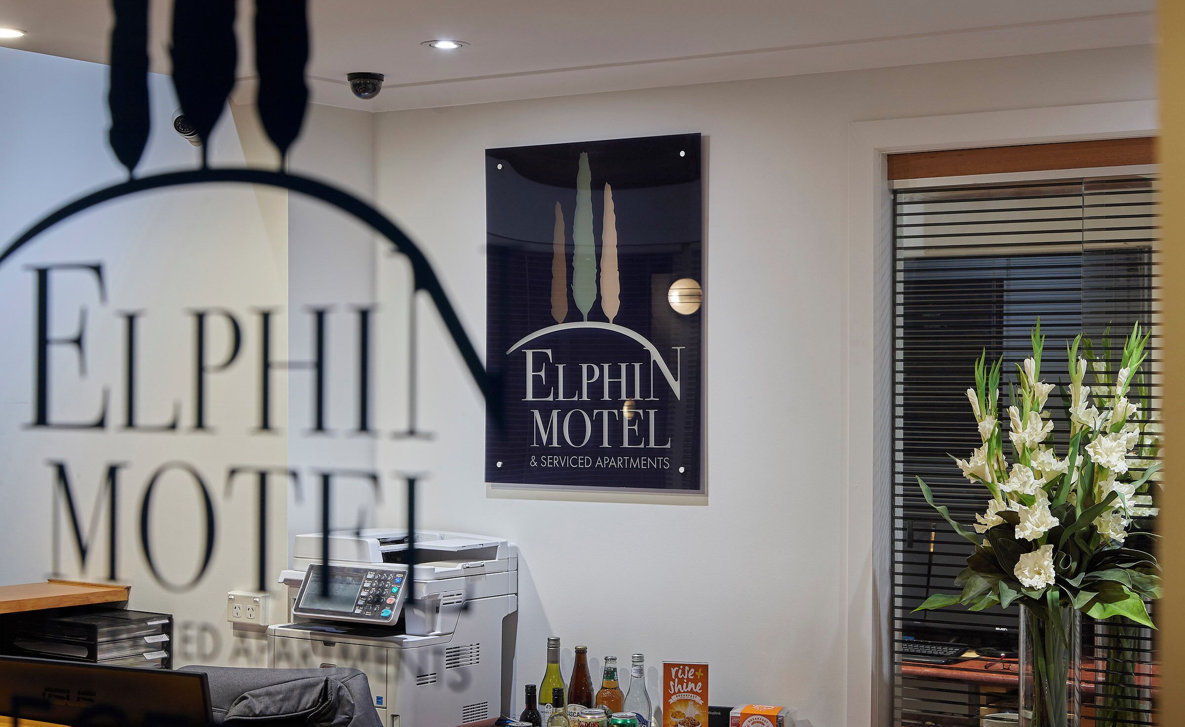 A reception desk with a coffee machine, a poster for Elphin Motel, and a window with blinds.
