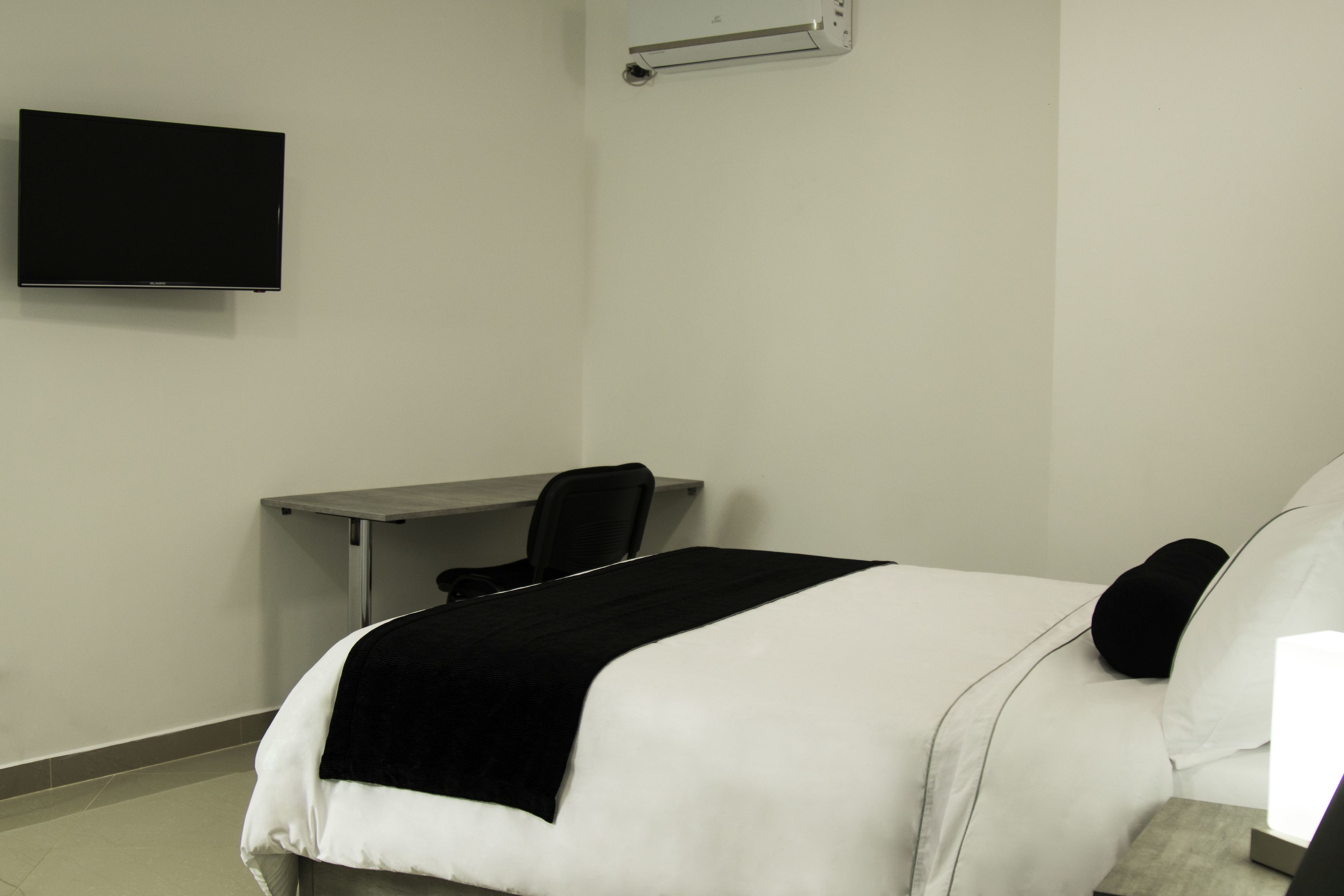 A hotel room with a bed, a desk, a chair, and a flat-screen TV mounted on the wall.