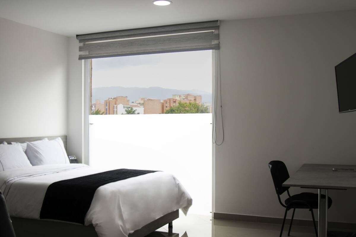 A hotel room with a bed, a desk, and a chair. There is a window with a view of buildings and mountains.