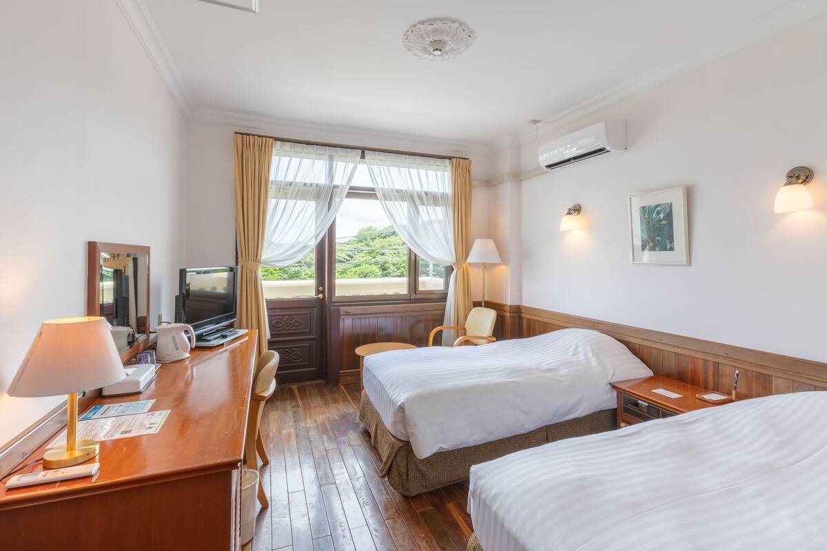 Hotellihuone, jossa on kaksi sänkyä, kirjoituspöytä lampun kanssa, televisio ja ikkuna, jossa on verhot.