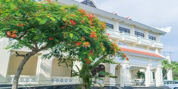 Un edificio de estilo colonial con balcón, ventanas de arco y un árbol con flores rojas en primer plano.