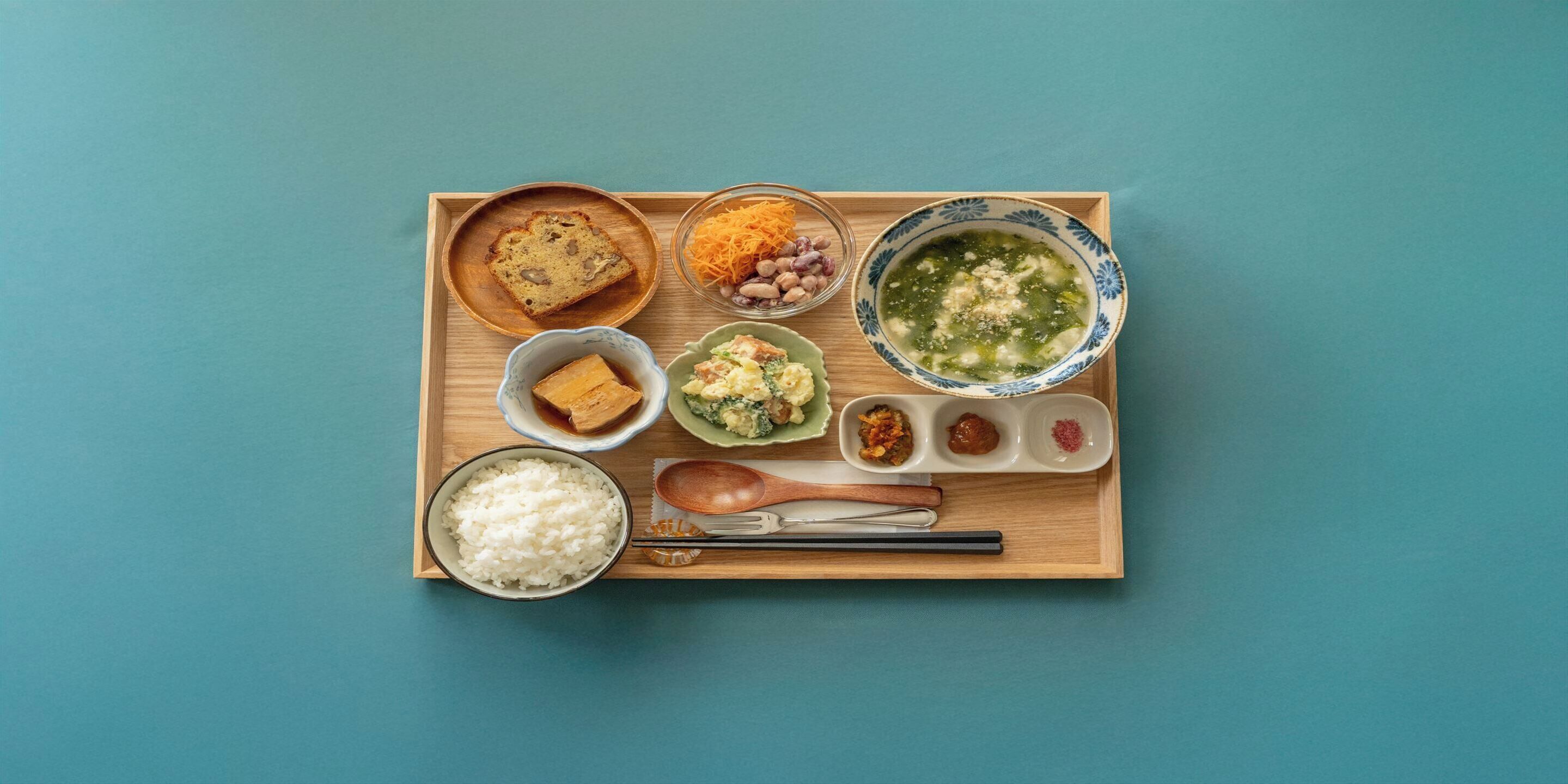 A wooden tray with various Japanese dishes including rice, tofu, and soup, arranged on a blue background.