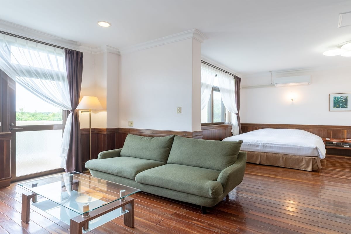 Hotellihuone, jossa on vihreä sohva, sänky, yöpöytä ja suuri ikkuna, jossa on verhot.