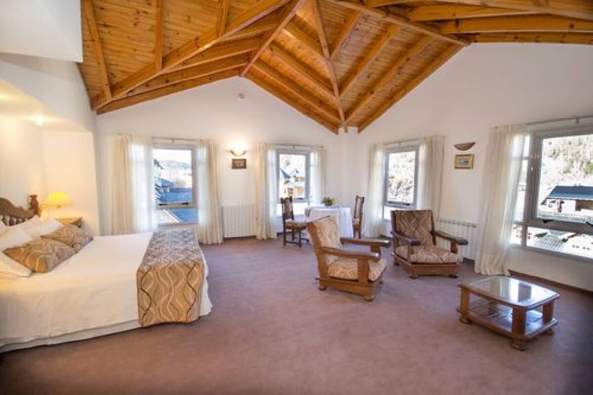 Ett hotellrum med ett träpanelat tak, en säng, två fåtöljer, ett litet bord och stora fönster med gardiner.