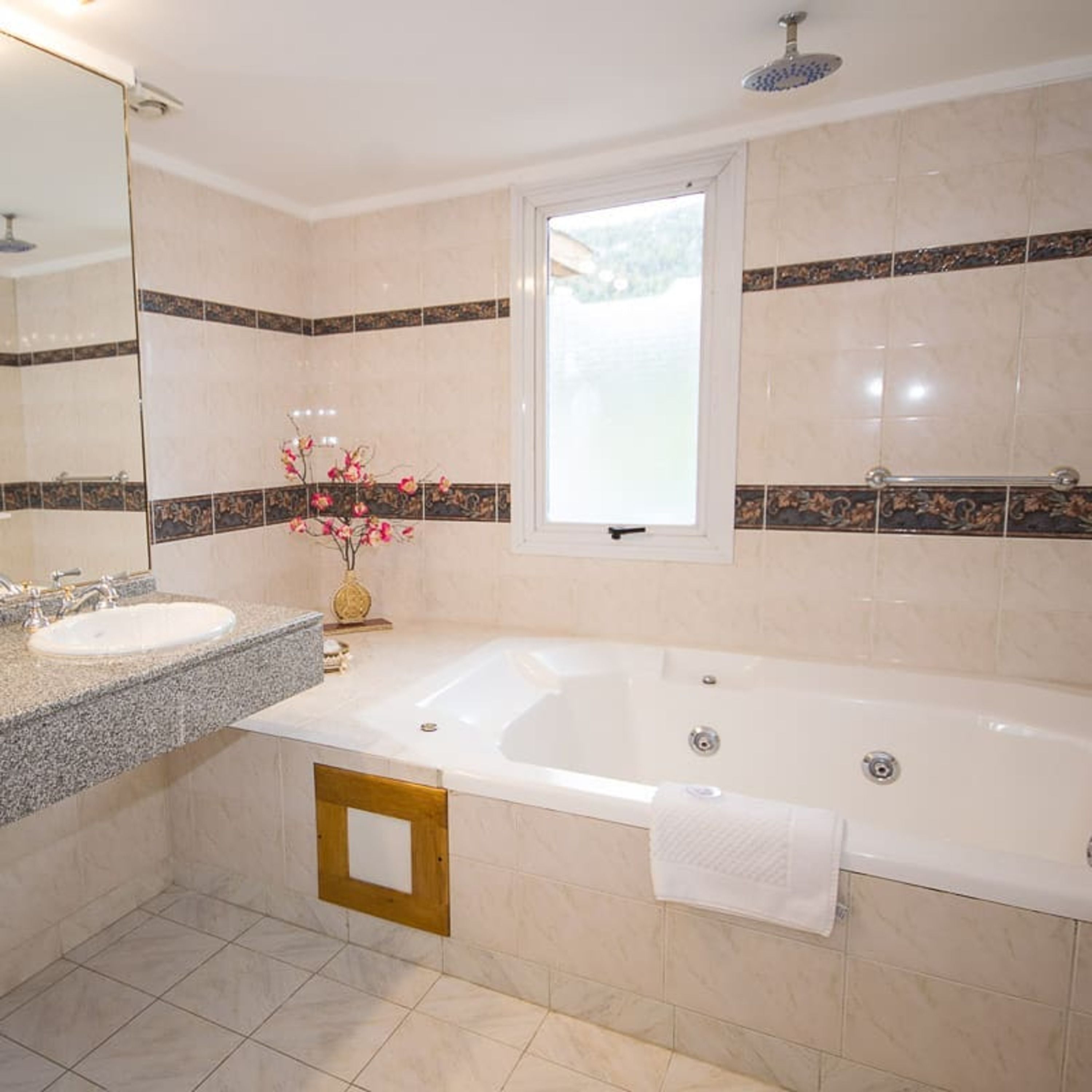 A bathroom with a large bathtub, a sink, and a mirror. There is a window above the bathtub and a decorative vase with flowers on the sink counter.
