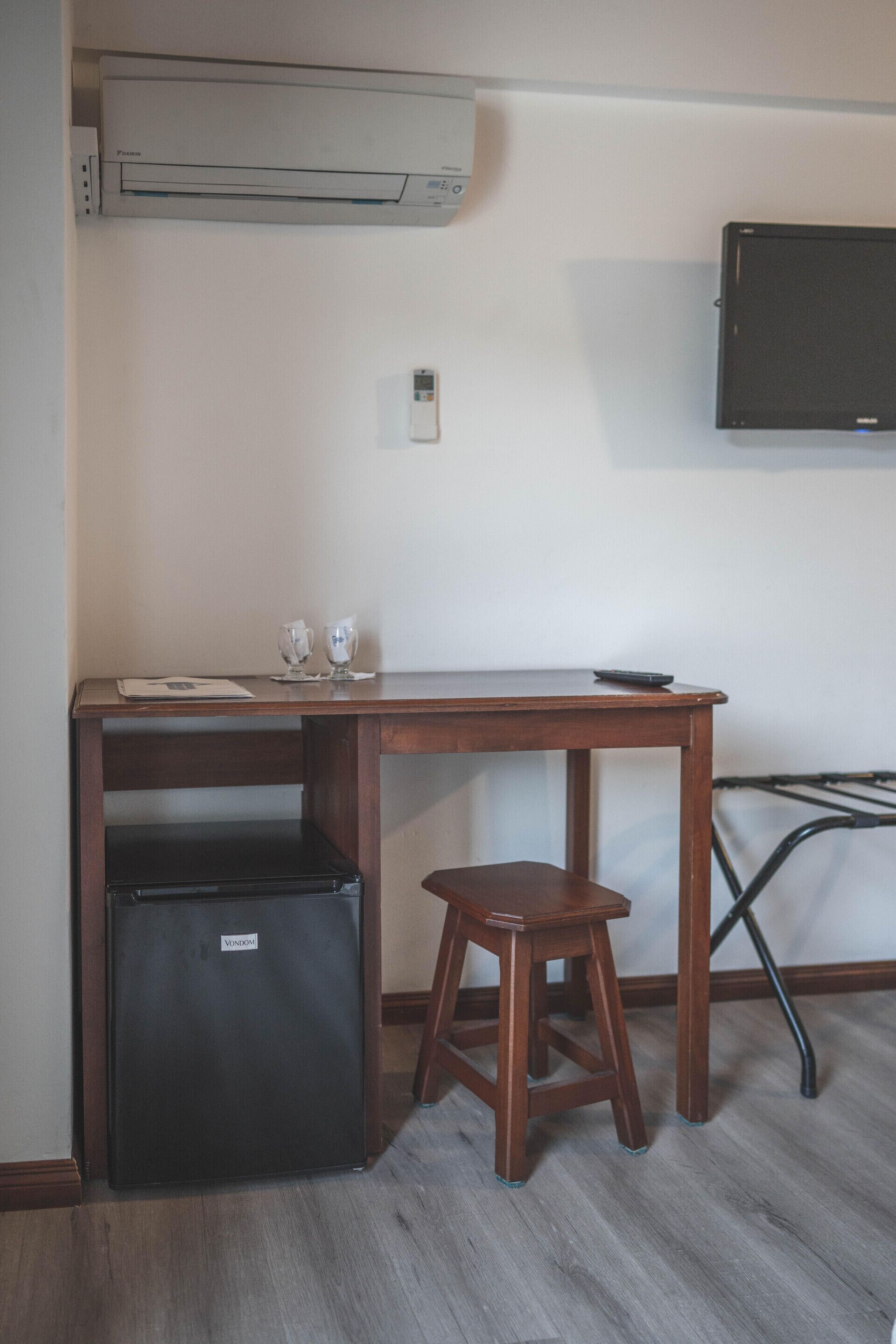 A compact room with a wooden desk, a small black refrigerator, a stool, a wall-mounted TV, and a wall-mounted air conditioner.