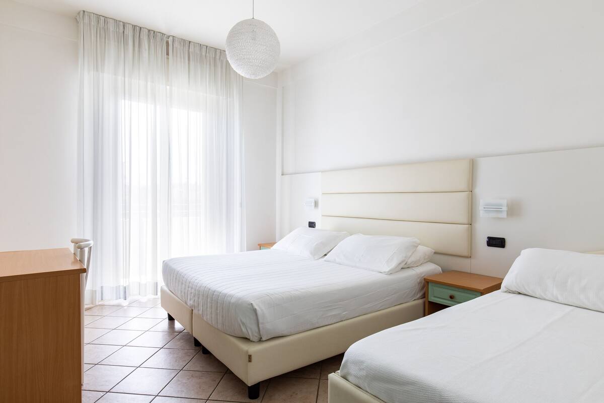 Una camera d'albergo con due letti, una scrivania in legno e un'ampia finestra con tende leggere.