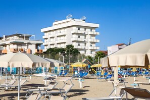A beach with sun loungers and umbrellas, a building with balconies, and a clear blue sky. - Hotel Palos (Rimini)
