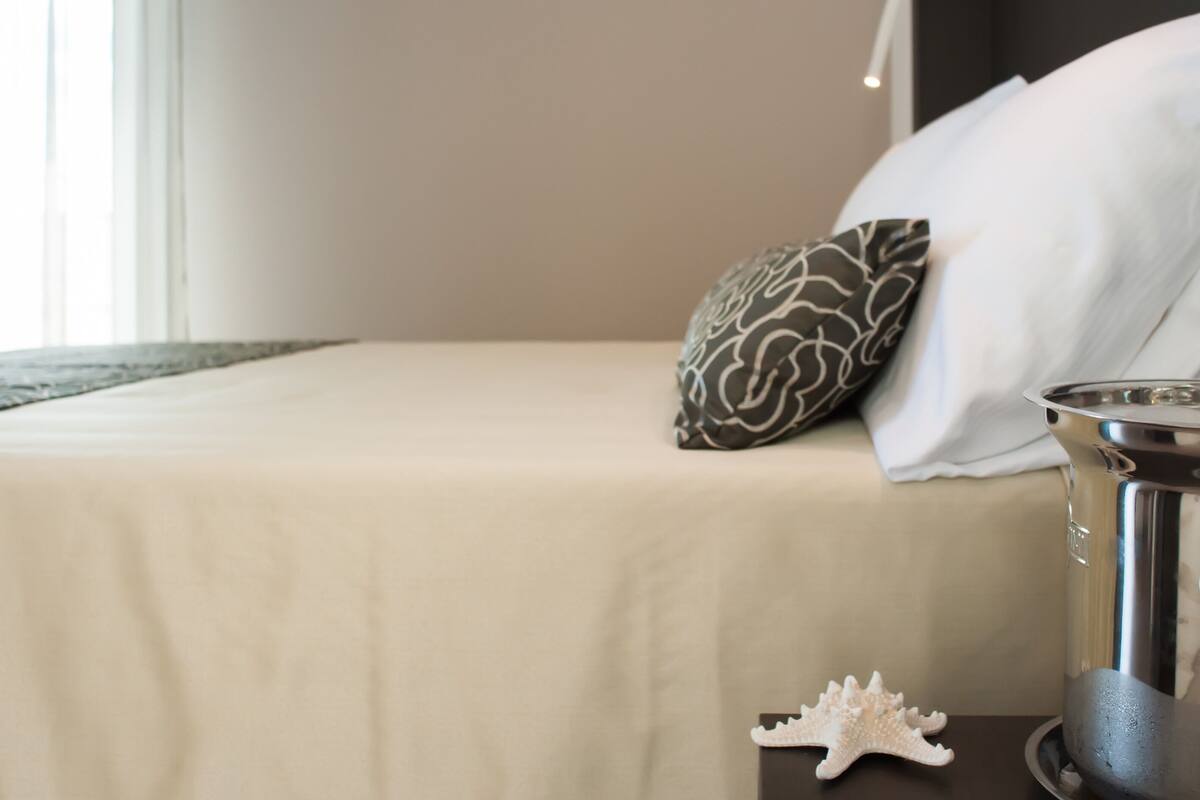 Un letto rifatto con cura, con una coperta beige e due cuscini, uno decorato e uno bianco liscio.